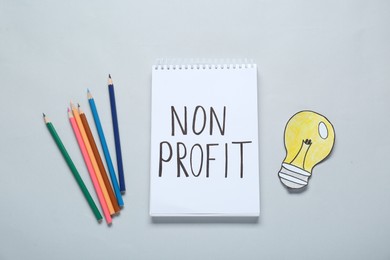Photo of Notebook with phrase Non Profit, pencils and paper lamp bulb on light background, flat lay