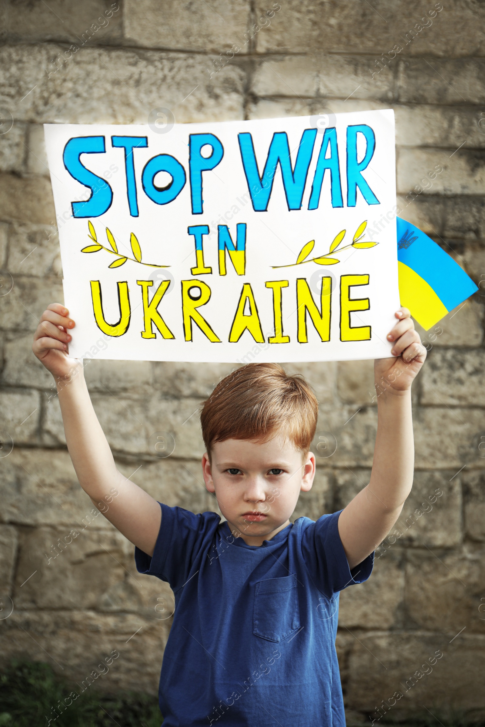 Photo of Sad boy holding poster Stop War In Ukraine and national flag against brick wall outdoors