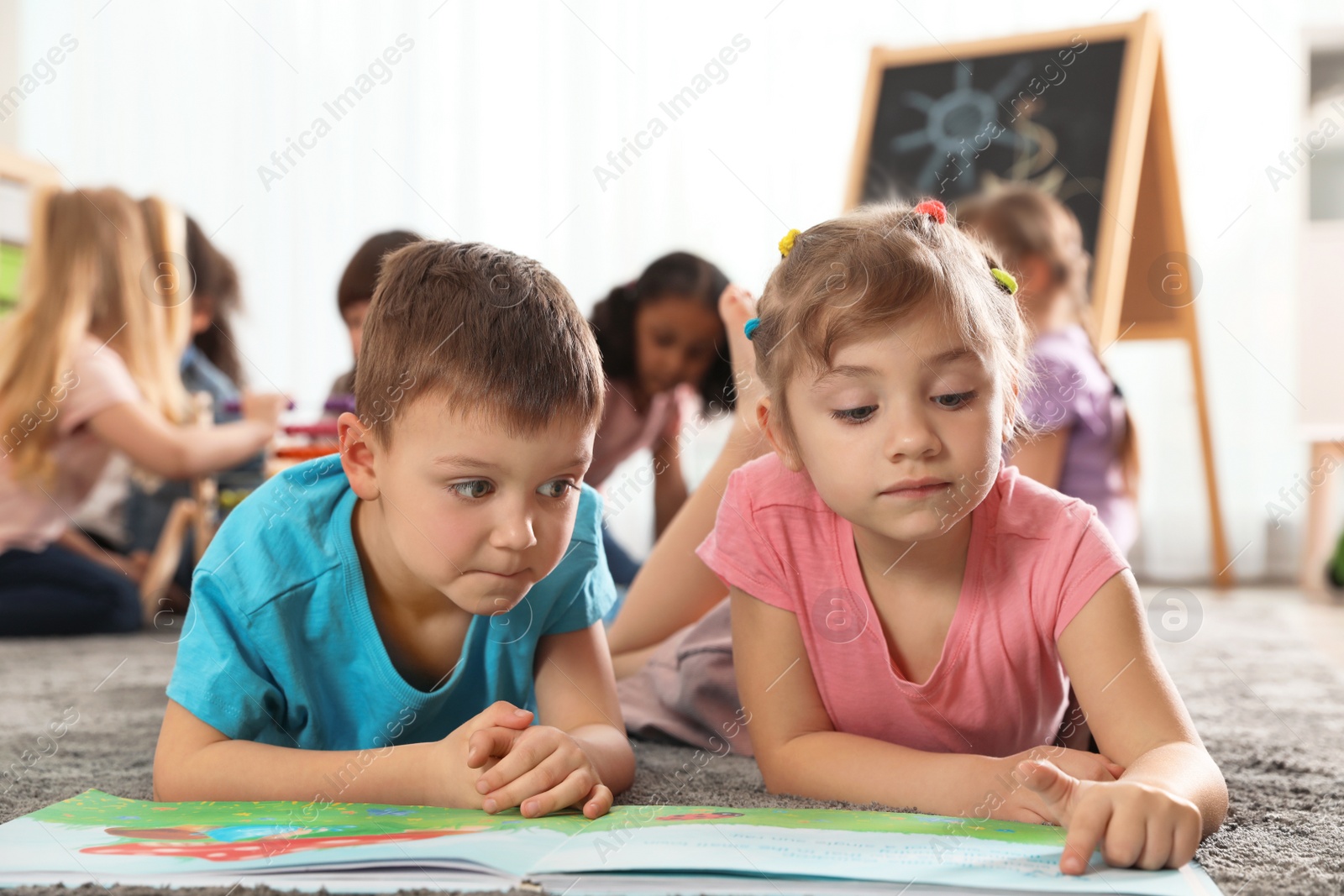 Photo of Cute kids reading book on floor while other children playing together in kindergarten