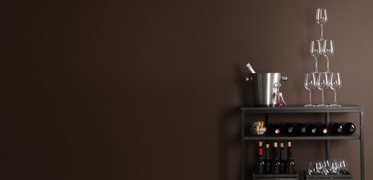 Photo of Rack with bottles of wine and glasses near brown wall, space for text