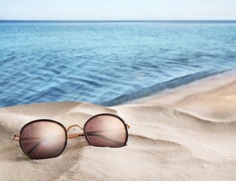 Stylish sunglasses on sandy beach near sea, space for text