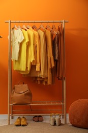 Rack with different stylish women`s clothes, shoes, backpack and pouf near orange wall indoors