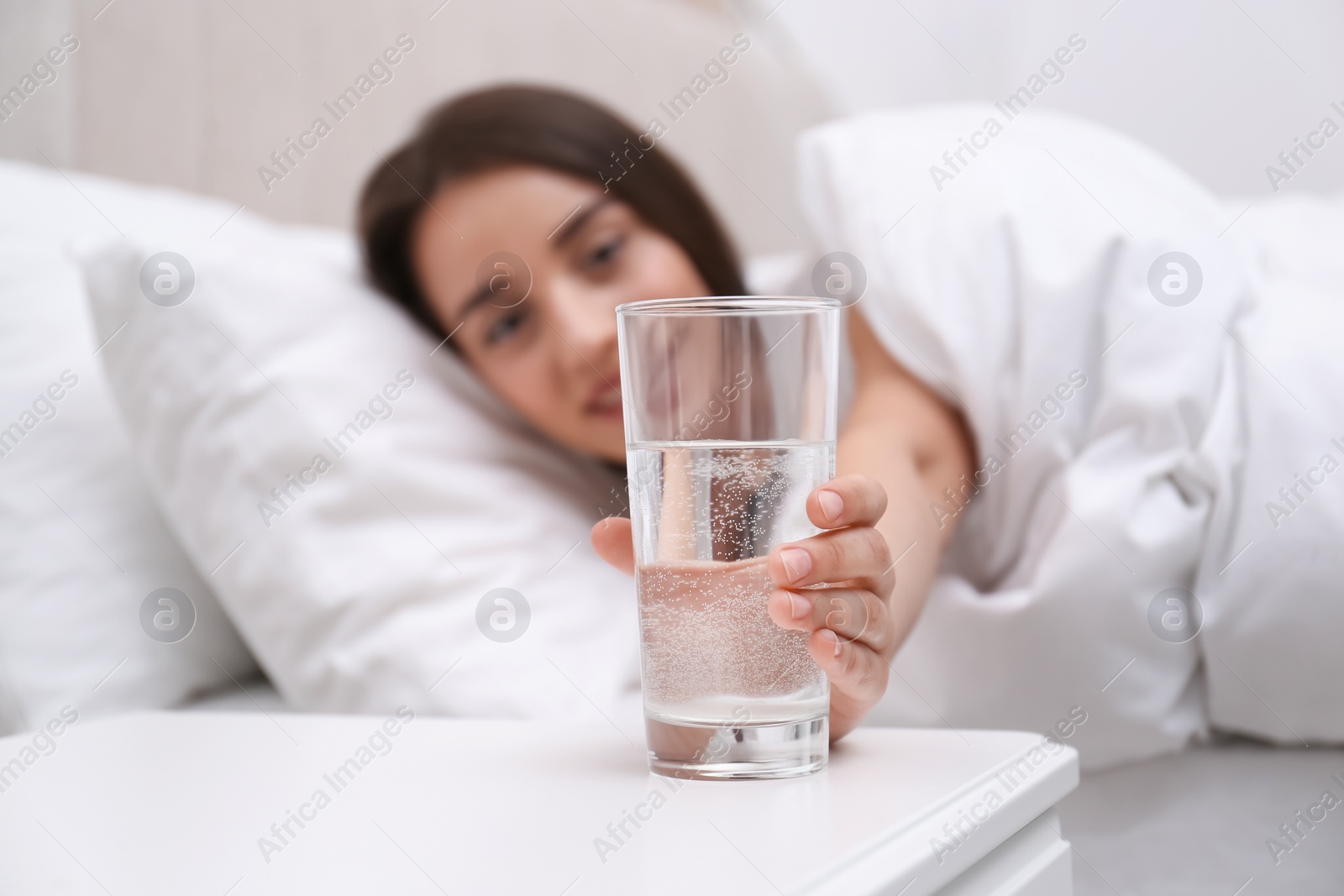 Photo of Young woman taking glass of water from nightstand at home, focus on hand