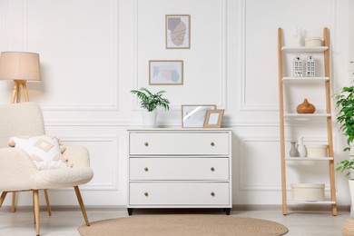 Photo of Picture frames hanging on white wall, chest of drawers and armchair in living room