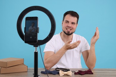 Photo of Smiling fashion blogger showing bow ties while recording video at table against light blue background