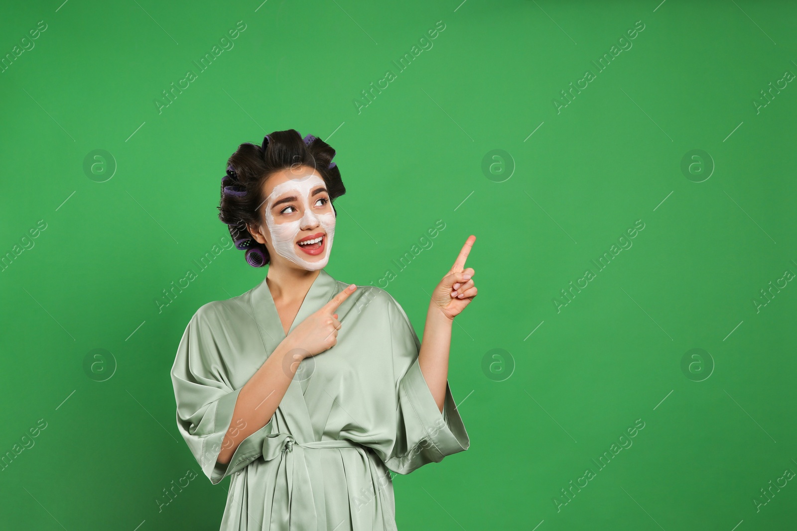 Photo of Happy young woman in silk bathrobe with hair curlers and facial mask on green background, space for text