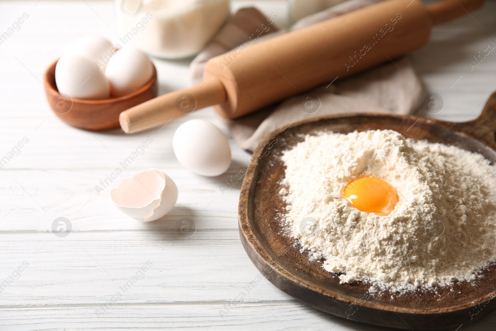 Photo of Making dough. Pile of flour with yolk, rolling pin and eggs on white wooden table, closeup