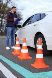Photo of Instructor near car with student during exam at driving school test track, focus on traffic cones