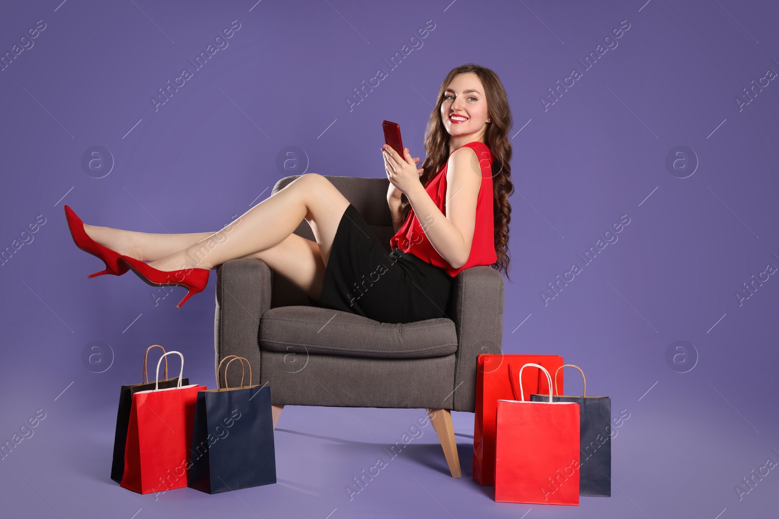 Photo of Happy woman resting on armchair with smartphone after shopping against purple background