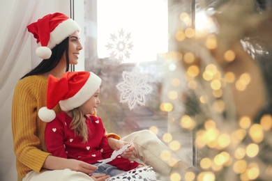 Photo of Mother and daughter in Santa hats with paper snowflakes near window at home