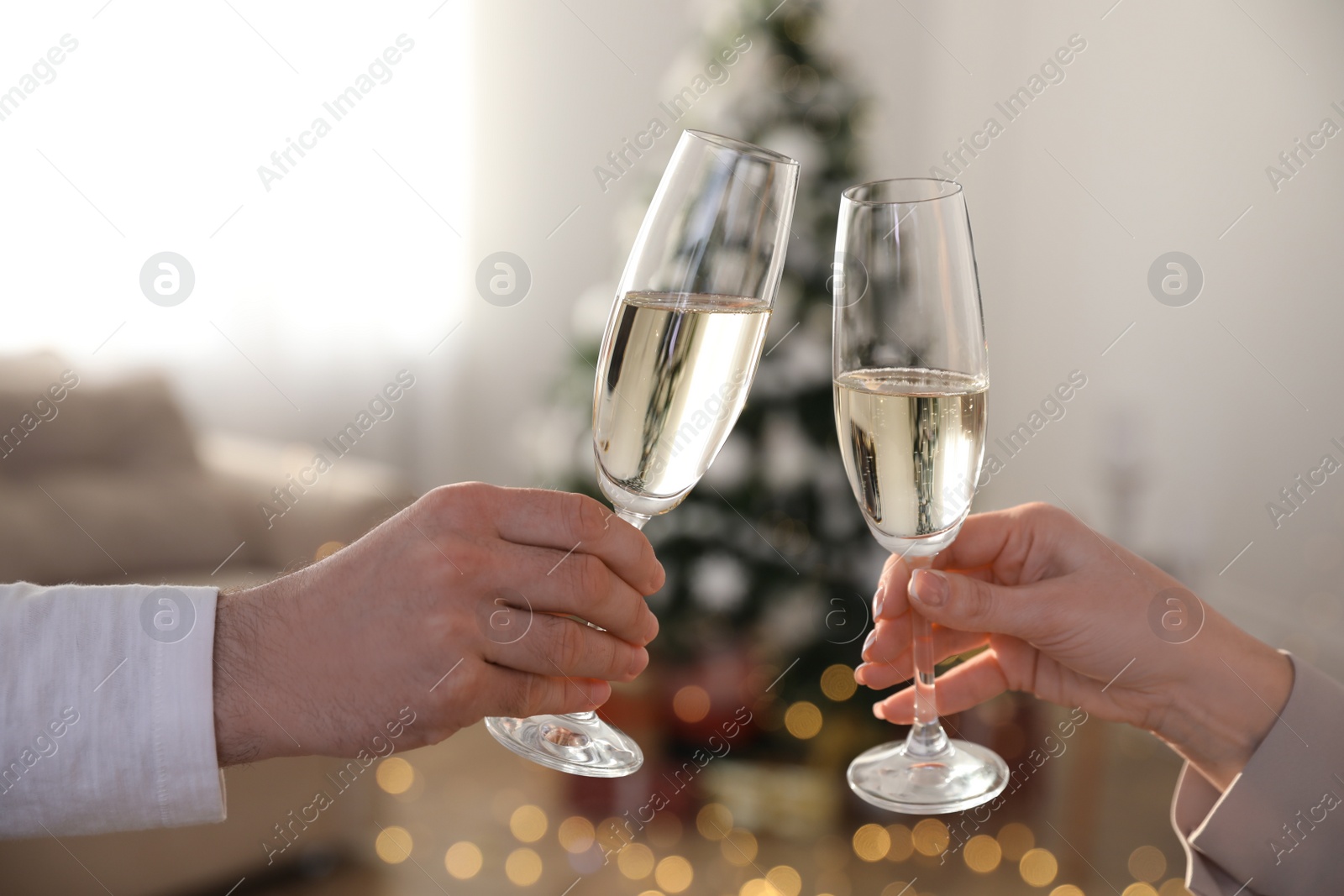 Photo of Couple clinking glasses of champagne in room decorated for Christmas, closeup. Holiday cheer and drink