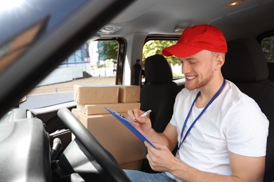 Young courier with clipboard and parcels in delivery car