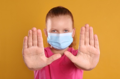 Photo of Little girl in protective mask showing stop gesture on yellow background. Prevent spreading of coronavirus