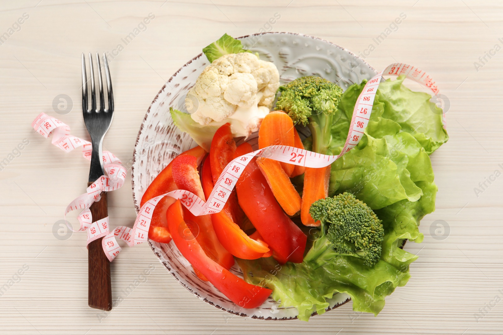 Photo of Healthy diet. Plate of fresh vegetables, measuring tape and fork on light wooden table, top view