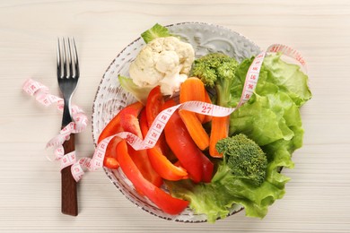 Photo of Healthy diet. Plate of fresh vegetables, measuring tape and fork on light wooden table, top view