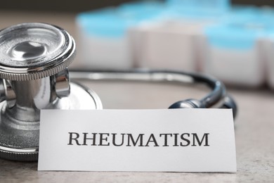 Photo of Card with word Rheumatism and stethoscope on light gray table, closeup