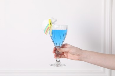 Woman holding glass of cocktail decorated with cotton candy and sour rainbow belt near white wall, closeup. Space for text