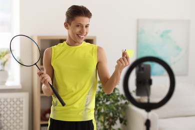 Photo of Smiling sports blogger holding badminton racket and shuttlecock while streaming online fitness lesson with smartphone at home