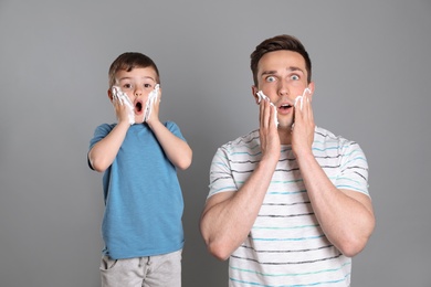 Photo of Emotional dad and his little son applying shaving foam on color background