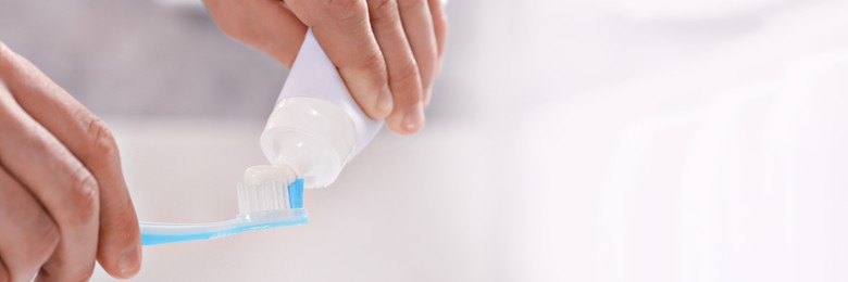 Man squeezing toothpaste from tube onto toothbrush in bathroom, closeup. Banner design with space for text