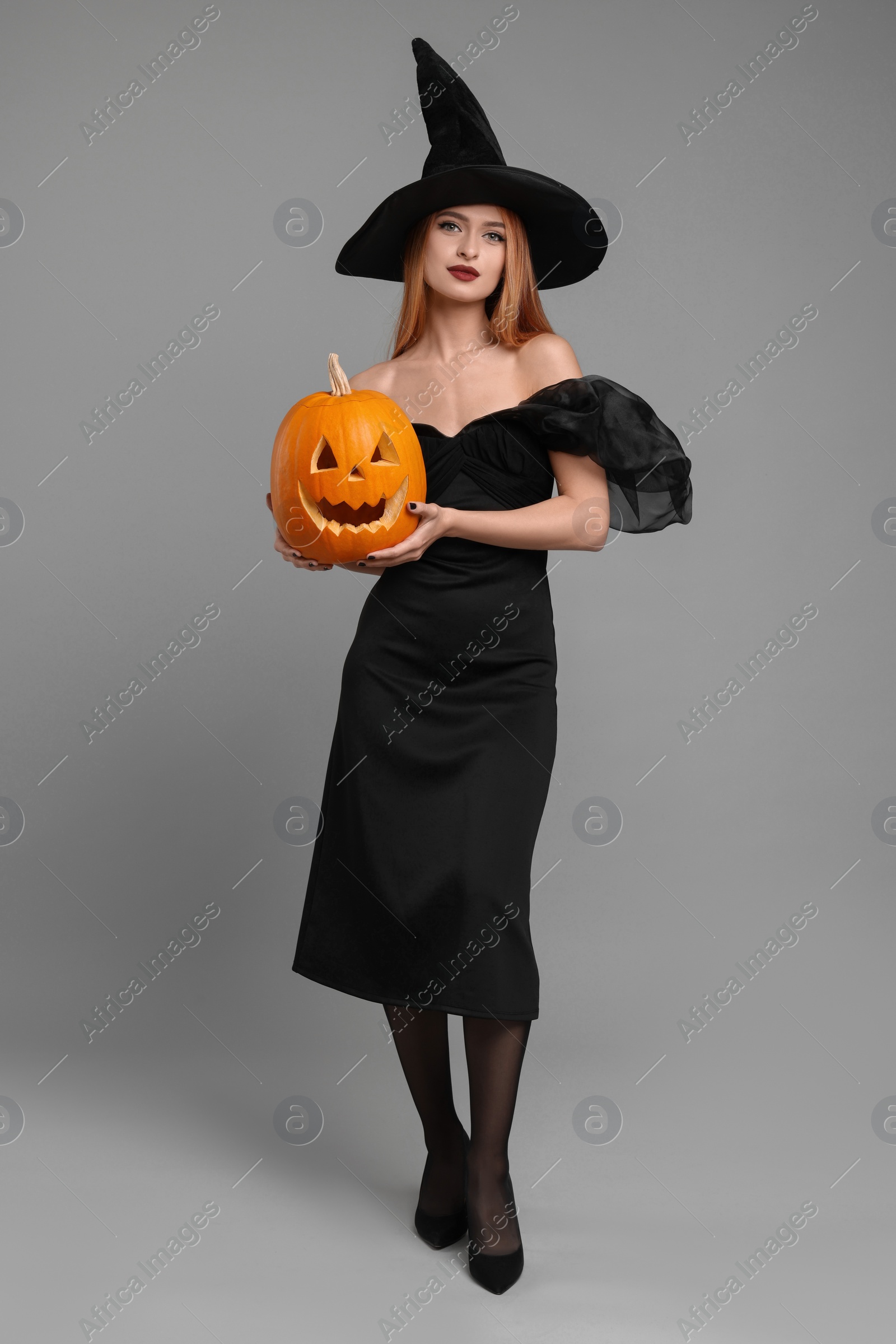 Photo of Young woman in scary witch costume with carved pumpkin on light grey background. Halloween celebration