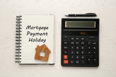 Photo of Notebook with words Mortgage Payment Holiday, house model, calculator and pen on white wooden table, flat lay