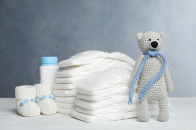 Photo of Baby diapers, booties, bottle and toy bear on white wooden table against grey background