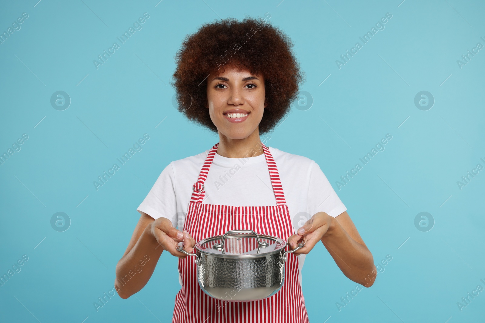 Photo of Happy young woman in apron holding cooking pot on light blue background