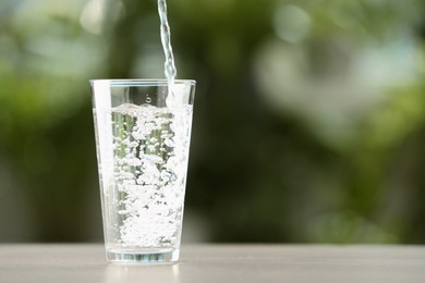Photo of Pouring water into glass on white table against blurred green background, closeup. Space for text