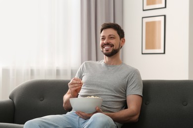 Happy man with bowl of popcorn watching movie via TV on sofa at home