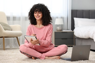 Beautiful young woman in stylish pyjama with laptop taking notes in notebook on floor at home