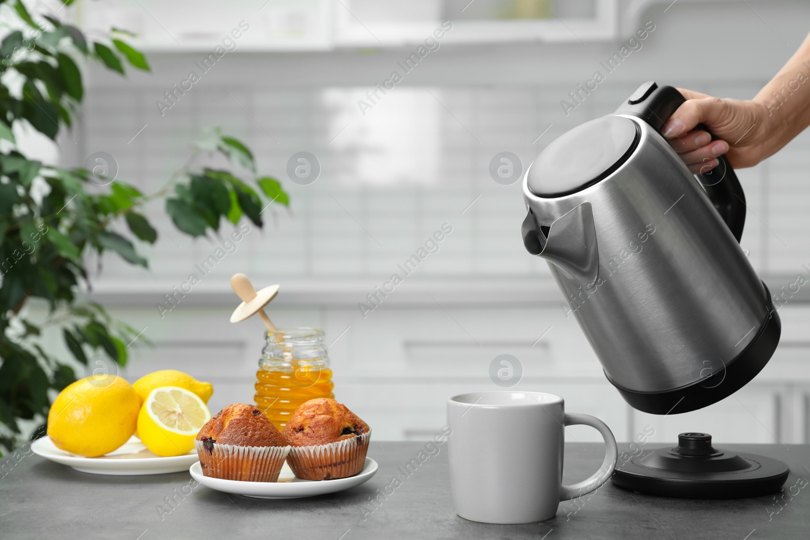 Photo of Woman pouring water from modern electric kettle into cup at grey table in kitchen, closeup