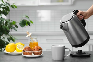 Photo of Woman pouring water from modern electric kettle into cup at grey table in kitchen, closeup
