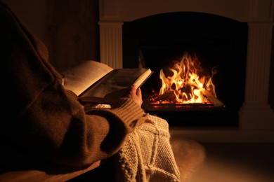 Woman reading book near fireplace indoors, closeup. Cozy atmosphere