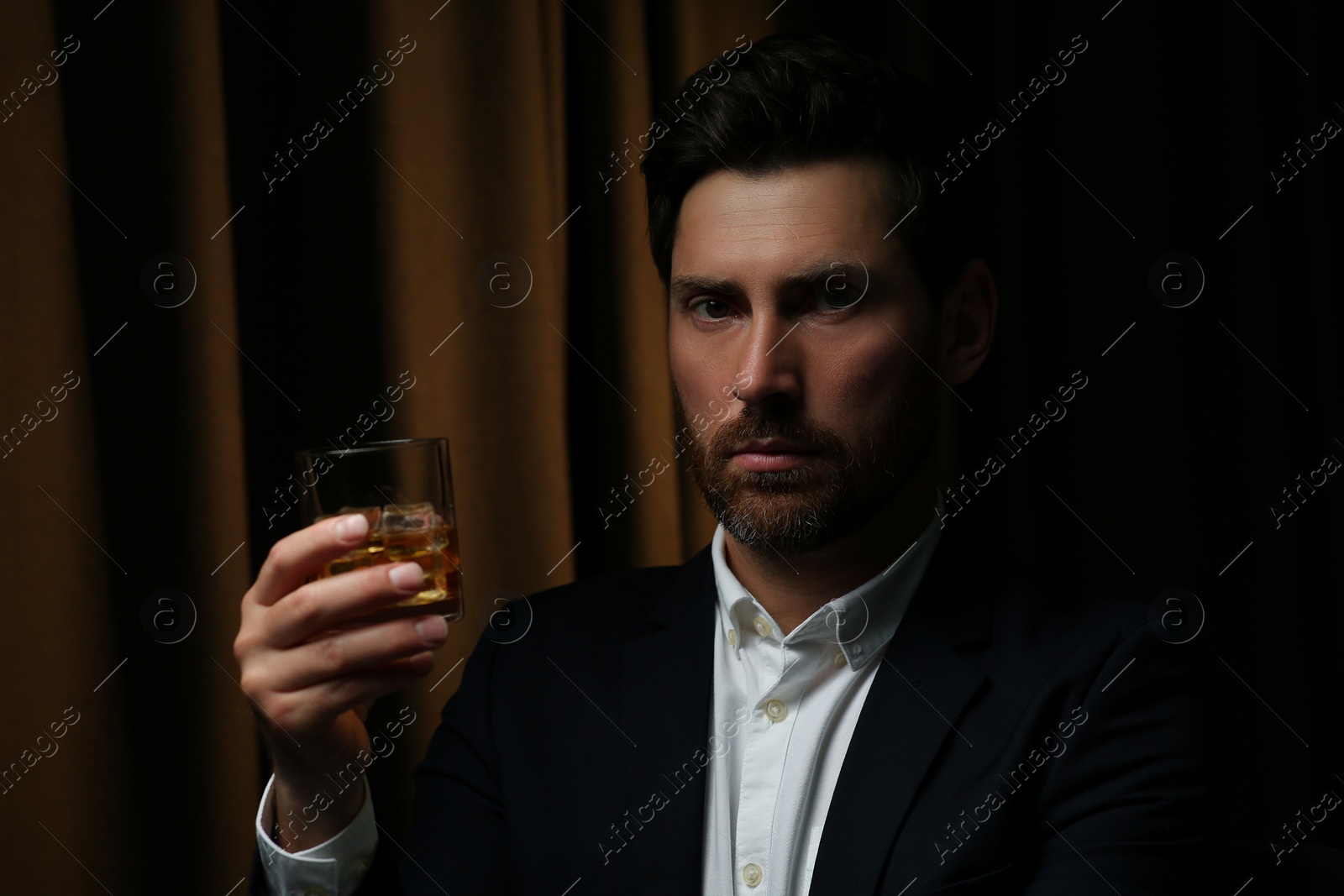 Photo of Man in suit holding glass of whiskey with ice cubes on brown background