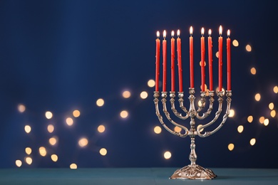Photo of Silver menorah with burning candles on table against blue background and blurred festive lights, space for text. Hanukkah celebration