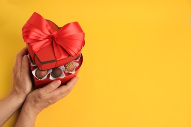 Woman with heart shaped box of delicious chocolate candies on yellow background, closeup. Space for text
