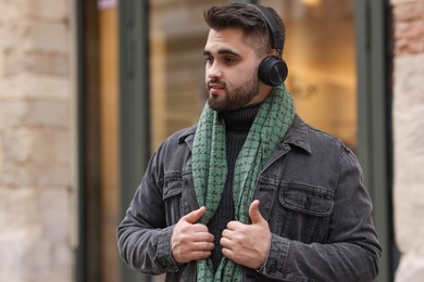 Photo of Handsome man in warm scarf and headphones near building outdoors. Space for text