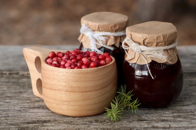 Photo of Tasty lingonberry jam in jars and cup with red berries on wooden table outdoors