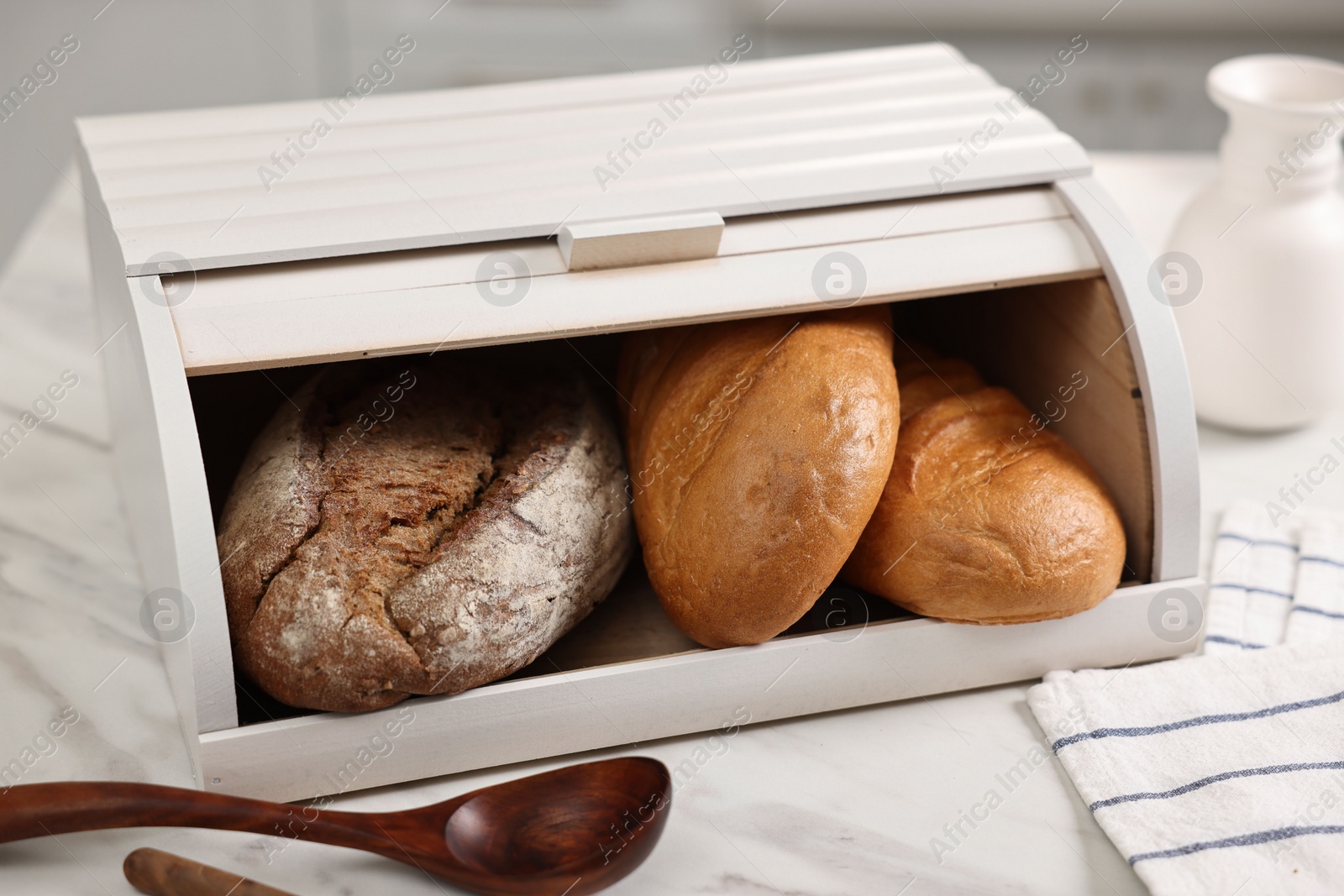 Photo of Wooden bread basket with freshly baked loaves and spoons on white marble table in kitchen