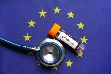 Photo of Stethoscope and test tube with blood sample on European Union flag background, flat lay. Coronavirus outbreak