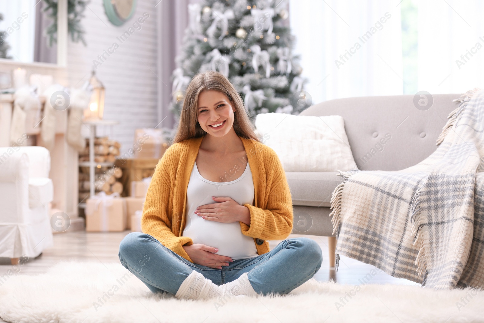 Photo of Young pregnant woman sitting on floor in room decorated for Christmas