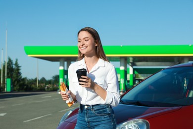 Photo of Beautiful young woman with coffee and hot dog near car at gas station