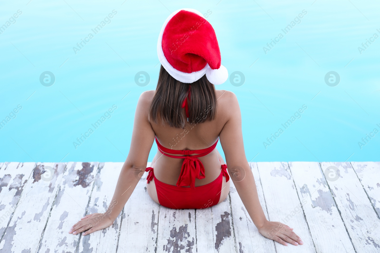 Photo of Young woman wearing Santa Claus hat near swimming pool, back view. Christmas vacation