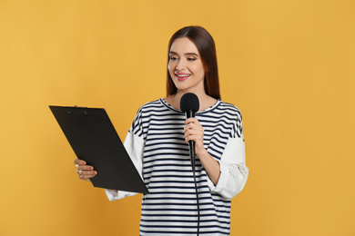 Photo of Young female journalist with microphone and clipboard on yellow background