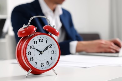 Red alarm clock and man working at table in office, closeup with space for text. Deadline concept