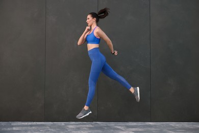 Beautiful woman in stylish sportswear jumping near black wall outdoors