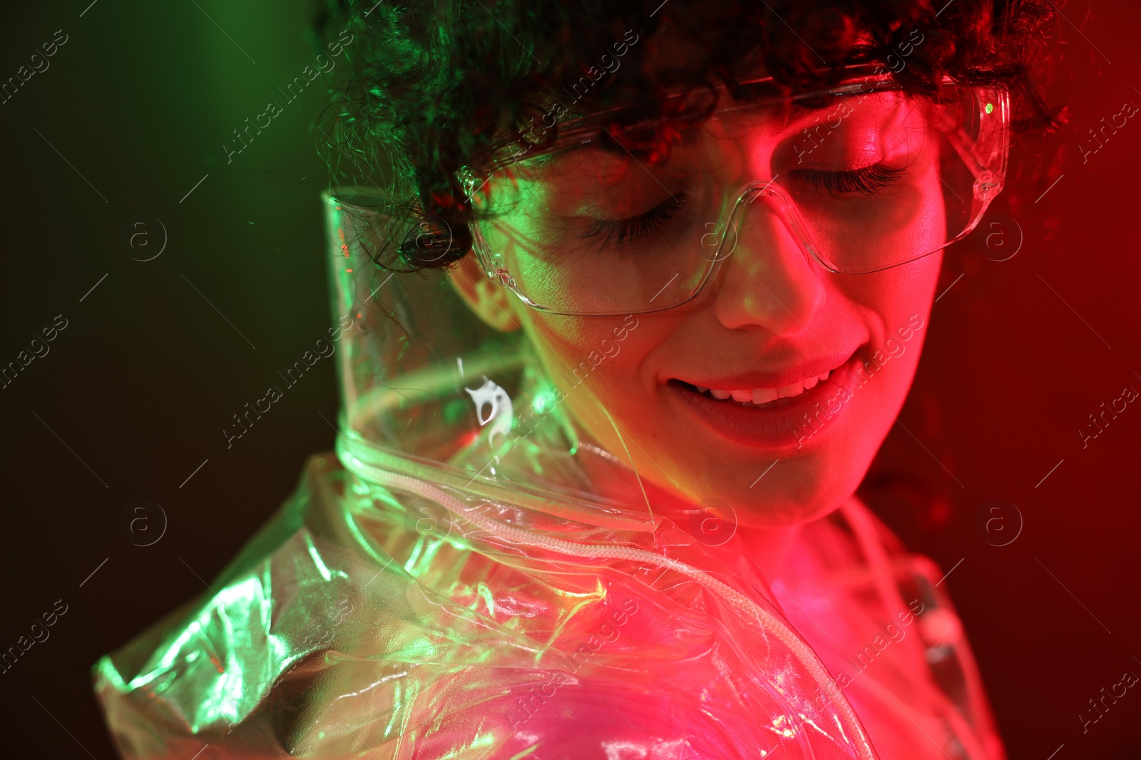 Photo of Beautiful young woman in transparent coat and sunglasses posing on color background in neon lights, closeup