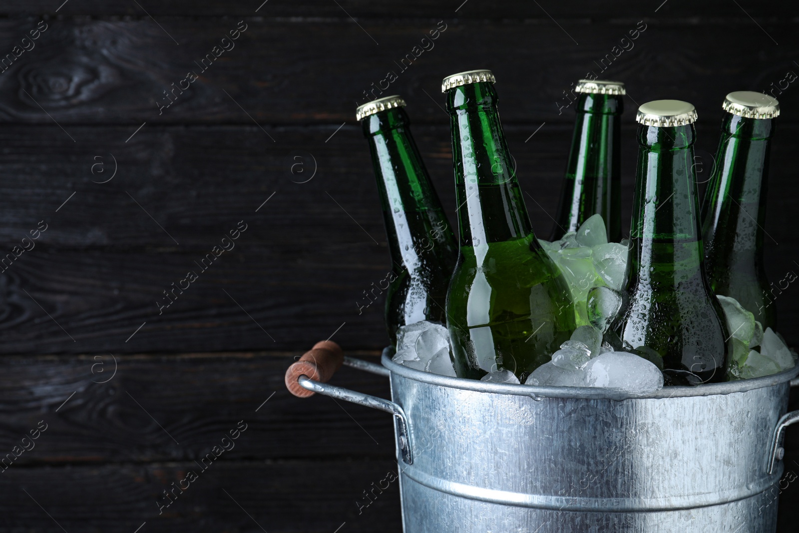 Photo of Metal bucket with bottles of beer and ice cubes on dark wooden background, closeup. Space for text