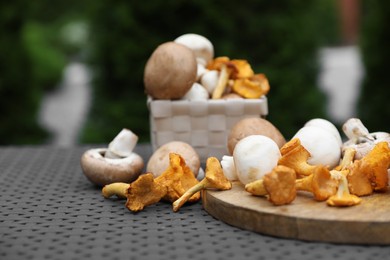 Photo of Different fresh mushrooms, board and basket on grey rattan table outdoors. Space for text